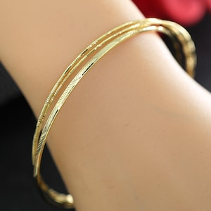 May include: Three gold bangles, each with a detailed, textured pattern. The bangles are displayed on a wrist, highlighting their shiny, reflective surface. The jewellery is a warm gold colour, with a blurred background.