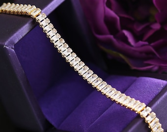 Puede incluir: Un brazalete de cadena de oro con piedras rectangulares transparentes. El brazalete está en una caja de terciopelo morado.