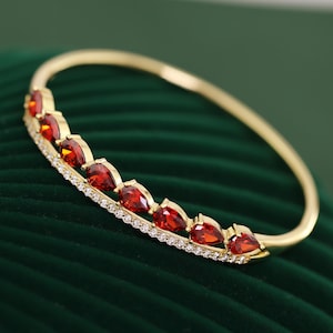 May include: A gold bangle bracelet with a row of red, teardrop-shaped gemstones. The gemstones are set along a band of small, clear crystals. The bracelet is displayed on a dark green, textured surface.