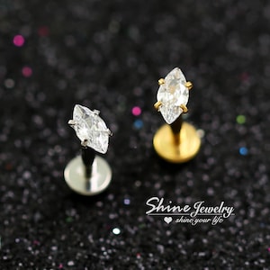 以下が含まれることがあります： マーキス型のクリアクリスタルラブレットスタッド2つ。1つはシルバーのポストとベース、もう1つはゴールドのポストとベースです。ジュエリーは暗くキラキラした背景にセットされています。「Shine Jewelry」の文字が見えます。
