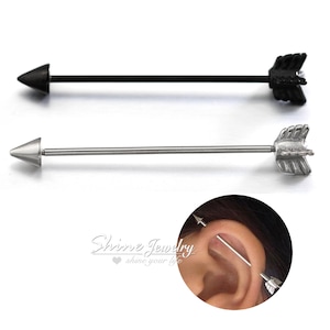 May include: Two industrial barbell earrings shaped like arrows. One is black with a black arrowhead and fletching. The other is silver with a silver arrowhead and fletching. The earrings are shown in an ear piercing.