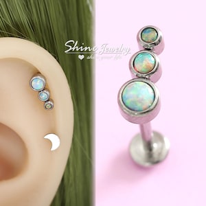 May include: A silver-colored ear piercing with three opal stones of varying sizes. The piercing is shown next to an ear with three opal studs and a crescent moon stud. The text "Shine Jewelry" is visible.