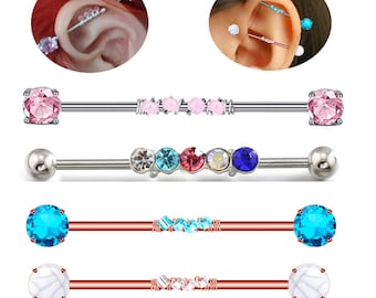 May include: A collection of industrial barbell earrings in various designs. The earrings feature different colored gemstones, including pink, blue, and clear stones. Some have a row of small gems, while others have larger, round stones at each end. The metal is silver and copper.