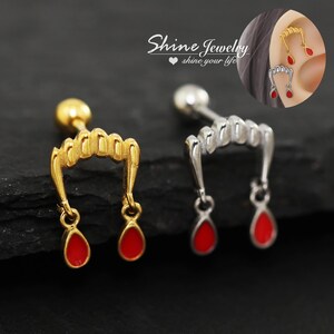 May include: Two vampire fang stud earrings, one gold-toned and one silver-toned, each featuring a row of teeth and two dangling red teardrop-shaped charms. The earrings have a ball-end post back and are shown in an inset image worn on an ear.