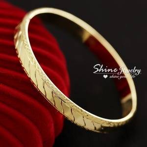 May include: A gold bangle bracelet with a textured, diamond-shaped pattern. The bracelet is on a red velvet background.