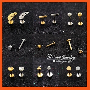 May include: A collection of lip and ear studs in gold and silver tones. Designs include feathers, bars, spheres, hearts, and clusters of small spheres. The studs are displayed on a dark, glittery surface. The text "Shine Jewelry shine your life" is visible.