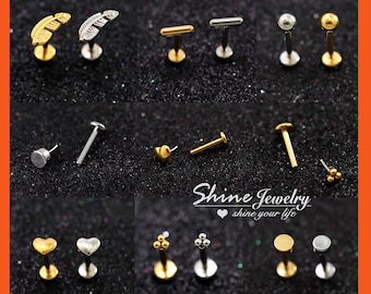 May include: An assortment of gold and silver lip and ear studs. The designs include feathers, bars, spheres, hearts, and clusters of small spheres. The studs are presented on a dark, glittery surface. The text "Shine Jewelry shine your life" is visible.