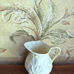 May include: Cream-colored ceramic pitcher with a detailed floral relief pattern. The pitcher has a curved handle and a scalloped rim. The background features a floral wallpaper design in muted tones of beige, brown, and green.