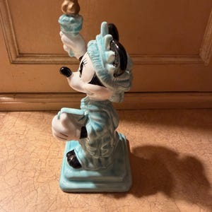 May include: A ceramic figurine of Minnie Mouse dressed as the Statue of Liberty. The figurine is primarily light blue with white gloves and a black face. She holds a golden torch aloft and stands on a square base.