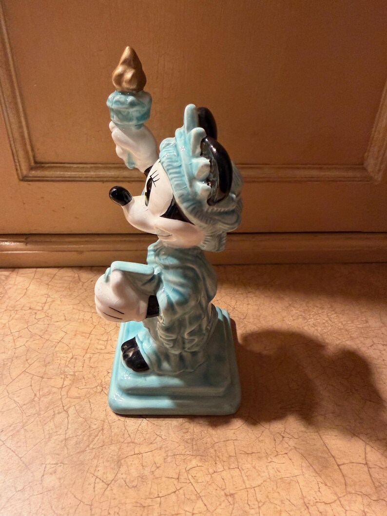 May include: A ceramic figurine of Minnie Mouse dressed as the Statue of Liberty. The figurine is primarily light blue with white gloves and a black face. She holds a golden torch aloft and stands on a square base.