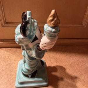 May include: A ceramic figurine of a character in a Statue of Liberty costume. The figure is primarily light blue with black accents and a gold torch. The figurine is standing on a square base.