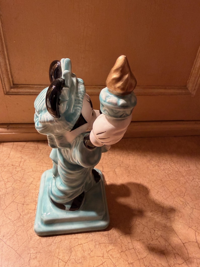 May include: A ceramic figurine of a character in a Statue of Liberty costume. The figure is primarily light blue with black accents and a gold torch. The figurine is standing on a square base.