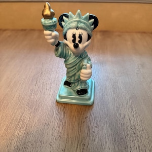 May include: A ceramic figurine of Minnie Mouse as the Statue of Liberty. The figurine is light blue with black accents and a gold torch. It stands on a square base and is a decorative item.