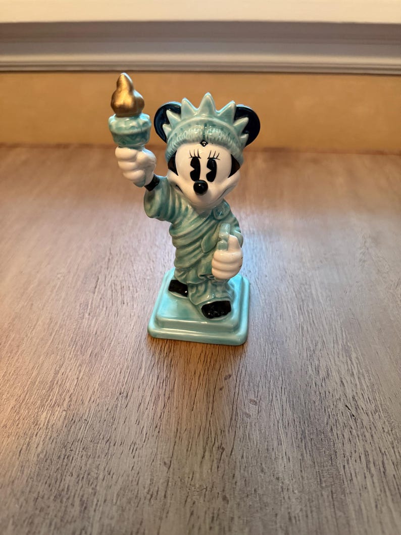 May include: A ceramic figurine of Minnie Mouse as the Statue of Liberty. The figurine is light blue with black accents and a gold torch. It stands on a square base and is a decorative item.