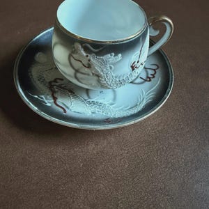 May include: A porcelain teacup and saucer set. The cup and saucer are decorated with a dragon motif in white and red against a black and gray background. The cup has a gold handle and rim.