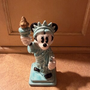 May include: A ceramic figurine of Minnie Mouse as the Statue of Liberty. The figurine is light blue with black ears and shoes, holding a gold torch. It stands on a square base and is a collectible item.