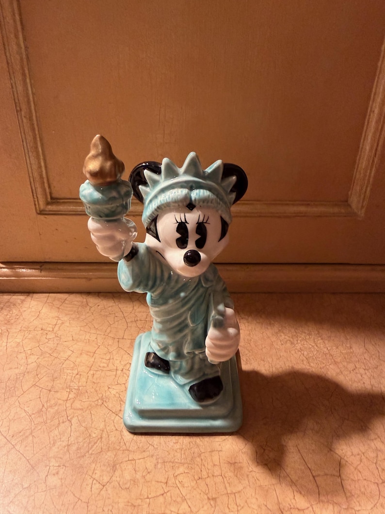 May include: A ceramic figurine of Minnie Mouse as the Statue of Liberty. The figurine is light blue with black ears and shoes, holding a gold torch. It stands on a square base and is a collectible item.