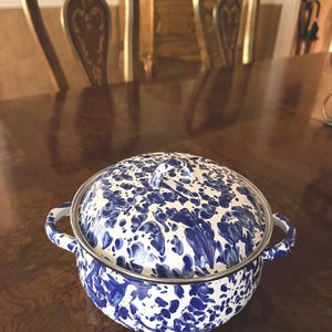 May include: A blue and white enamelware pot with a lid. The pot has a speckled pattern and two handles. The lid has a small knob. The pot is sitting on a wooden table.