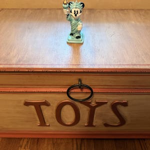 May include: A light blue ceramic figurine of Mickey Mouse as the Statue of Liberty, holding a torch. The figurine is on a wooden toy chest with the word "TOYS" in large letters.