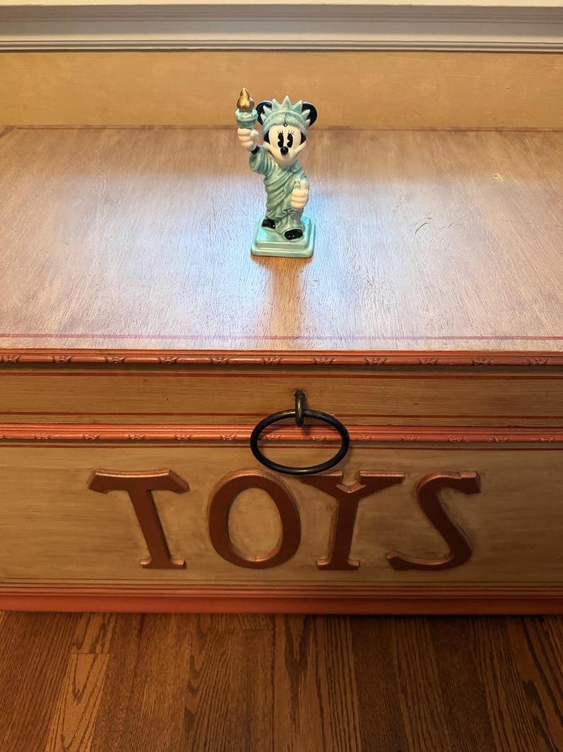 May include: A light blue ceramic figurine of Mickey Mouse as the Statue of Liberty, holding a torch. The figurine is on a wooden toy chest with the word "TOYS" in large letters.