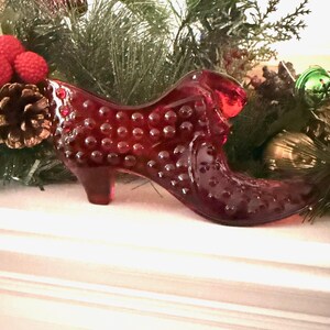 May include: A ruby red glass slipper with a textured surface, displayed with Christmas greenery and ornaments. The decorative shoe features a small heel and a classic slipper shape, perfect for holiday decor.