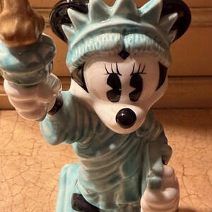 May include: A ceramic figurine of Minnie Mouse as the Statue of Liberty. The figurine is mainly light blue with black and white details. Minnie Mouse holds a golden torch and wears a crown.