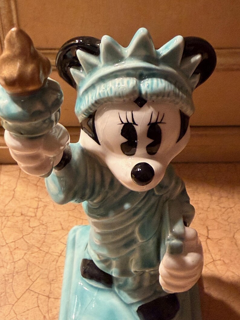 May include: A ceramic figurine of Minnie Mouse as the Statue of Liberty. The figurine is mainly light blue with black and white details. Minnie Mouse holds a golden torch and wears a crown.