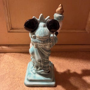 May include: A ceramic figurine of the Statue of Liberty, viewed from the back. The statue is light blue with black eyes and a torch. The base is square and the statue is standing on it. The torch has a gold flame.