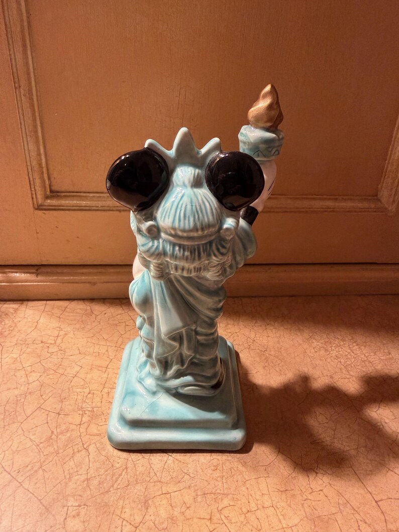 May include: A ceramic figurine of the Statue of Liberty, viewed from the back. The statue is light blue with black eyes and a torch. The base is square and the statue is standing on it. The torch has a gold flame.
