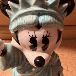 May include: A ceramic figurine of Minnie Mouse as the Statue of Liberty. The figurine has a light blue crown and robe, with black accents on the ears and eyes. The face is white with black eyes and a black nose.