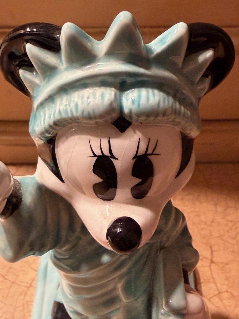 May include: A ceramic figurine of Minnie Mouse as the Statue of Liberty. The figurine has a light blue crown and robe, with black accents on the ears and eyes. The face is white with black eyes and a black nose.