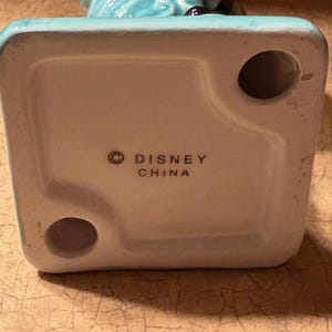 May include: A ceramic figurine in turquoise and white. The base is white with the text "&copy; DISNEY CHINA" printed in black. The figurine has a glossy finish and is likely a collectible item. The design is detailed and the colours are vibrant.