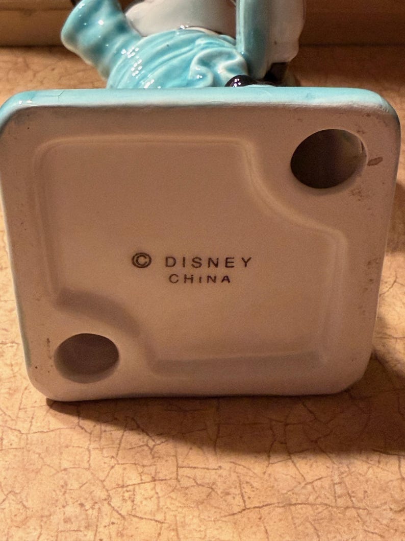 May include: A ceramic figurine in turquoise and white. The base is white with the text "&copy; DISNEY CHINA" printed in black. The figurine has a glossy finish and is likely a collectible item. The design is detailed and the colours are vibrant.