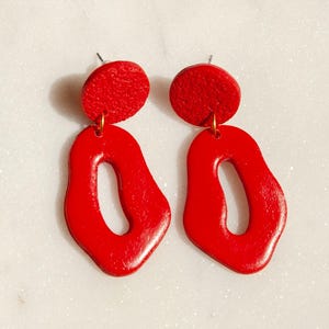 Könnte beinhalten: Ein Paar leuchtend rote Ohrringe. Jeder Ohrring hat einen kleinen, strukturierten, kreisförmigen Stecker, der mit einem größeren, unregelmäßigen, oval geformten Anhänger mit glatter, glänzender Oberfläche verbunden ist. Auf weißem Hintergrund.