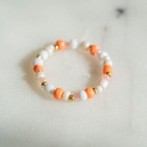 Könnte beinhalten: Ein Armband aus weißen und orangefarbenen Perlen mit goldenen Akzenten.