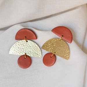 Könnte beinhalten: Ein Paar geometrische Ohrringe in einem Farbspiel aus Gold und Terrakotta. Die Ohrringe bestehen aus einem großen, gehämmerten goldenen Halbkreis, einem kleineren terrakottafarbenen Halbkreis und einer runden terrakottafarbenen Perle.