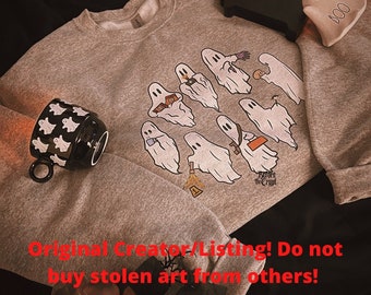 May include: A gray sweatshirt with a graphic of a line of ghosts holding various objects. The text "Before the Crypt" is printed below the ghosts. The sweatshirt is laying on a surface with a black mug with a ghost pattern and a white ceramic ghost figurine.