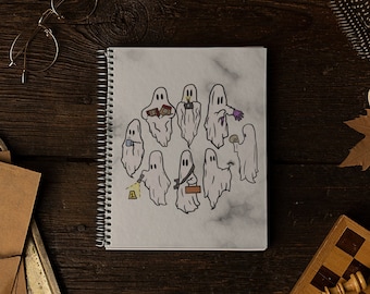 May include: A white spiral bound notebook with a drawing of ghosts holding various objects on a marble background. The notebook is on a dark brown wooden surface with other objects around it.