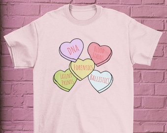 May include: A pink t-shirt with a graphic of five candy hearts. Each heart has a different forensic term written on it: DNA, Blood Stains, Forensics, Latent Prints, and Ballistics.