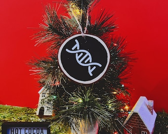 May include: A small artificial Christmas tree decorated with a black and white DNA ornament. The tree is surrounded by a gingerbread house, a gingerbread man, pine cones, and a sign that reads "HOT COCOA".