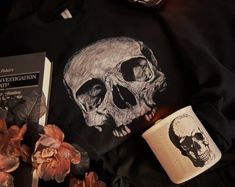 May include: A black sweatshirt with a white skull graphic print. Two candles with skull designs are on the sweatshirt, one is lit. A book titled "Fisher's Investigation of Death" is on the left side of the image.  Pink flowers are on the bottom left side of the image.