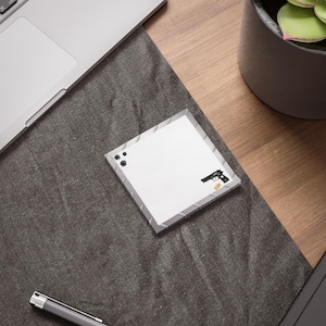 May include: Square white notepad with a gray border, featuring a black handgun graphic and bullet hole illustrations. A small, rectangular yellow graphic is next to the gun. The notepad is on a dark gray surface.