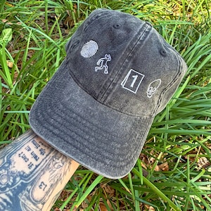 Op de afbeelding: Donkergrijze baseballcap met witte geborduurde ontwerpen: een vingerafdruk, een rennende figuur, het nummer 1 en een schedel. De cap, gemaakt van versleten stof, heeft een gebogen klep. Ligt op groen gras.