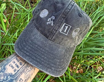 May include: A dark gray baseball cap with white embroidered designs, including a fingerprint, running figure, number 1, and a skull. The cap is made of a worn fabric and has a curved brim. The cap is sitting on a bed of green grass.