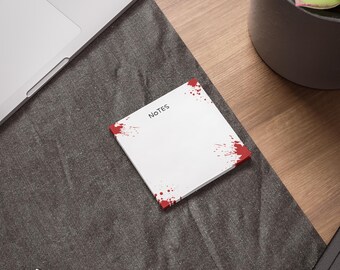 May include: A white square notepad with red splatter designs on the corners and edges. The word "NOTES" is printed in black at the top. The notepad is on a dark gray surface, near a laptop and a pen.