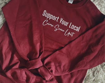 May include: A maroon sweatshirt with the text "Support Your Local Crime Scene Unit" in white. The sweatshirt has a crew neck and long sleeves with ribbed cuffs. The fabric appears soft and comfortable.