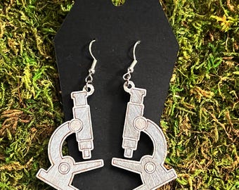 May include: A pair of silver microscope earrings. The earrings are made of wood and have a whitewash finish. The microscopes are detailed with a lens, stage, and base.