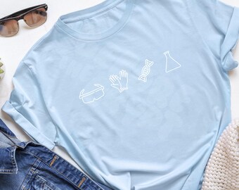 May include: Light blue t-shirt with white line art of science-related icons: goggles, gloves, DNA strand, and a flask. The shirt is folded, with sunglasses and a denim jacket visible in the background.