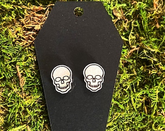 May include: A pair of wooden skull stud earrings. The earrings are white and have a black outline. They are displayed on a black coffin-shaped card.