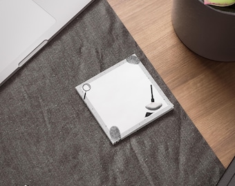May include: Square white notepad with a gray border and detective-themed illustrations, including a magnifying glass and a fingerprint. The notepad is on a dark gray surface, near a laptop and a pen.